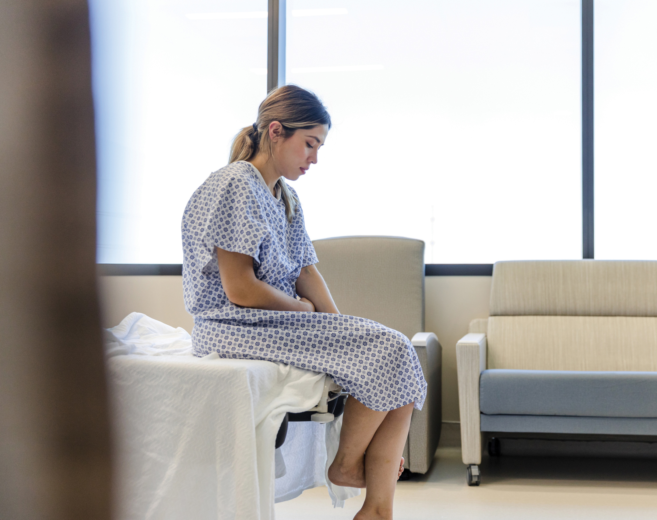 Anxious, sad, young woman wearing hospital gown looks down